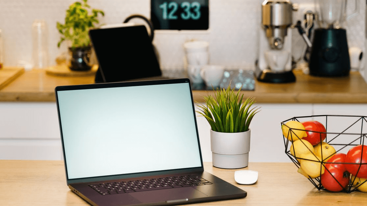 Laptop on a kitchen table beside a potted plant and a bowl of fruit